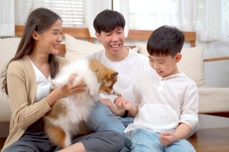 Happy cheerful Asian family father mother and son enjoy playing with a lovely little dog in living room together. Asian little boy staying with parent in living room with a pet.の写真素材