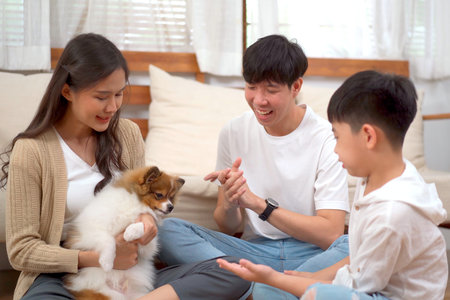 Happy cheerful Asian family father mother and son enjoy playing with a lovely little dog in living room together. Asian little boy staying with parent in living room with a pet.の写真素材