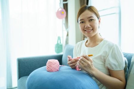 Happy cheerful Asian woman doing a crochet in living room in free time. Skillful Asian craftswoman knitting a handcraft crochet sweater and hat in cozy living room.の写真素材