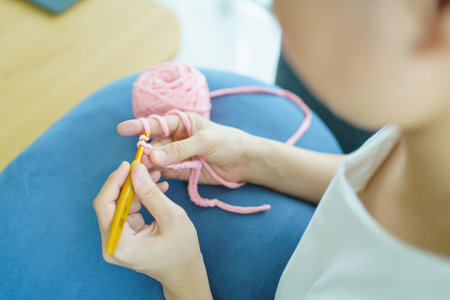 Happy cheerful Asian woman doing a crochet in living room in free time. Skillful Asian craftswoman knitting a handcraft crochet sweater and hat in cozy living room.の写真素材