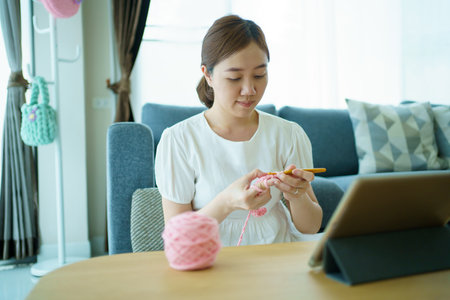 Happy cheerful Asian woman doing a crochet in living room in free time. Skillful Asian craftswoman knitting a handcraft crochet sweater and hat in cozy living room.の写真素材