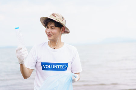 Asian young man picking up a plastic bottle garbage on the beach, male volunteer walking around the beach and collecting - cleaning a plastic waste.の写真素材
