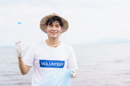 Asian young man picking up a plastic bottle garbage on the beach, male volunteer walking around the beach and collecting - cleaning a plastic waste.の写真素材