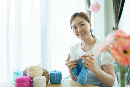 Happy cheerful Asian woman doing a crochet in living room in free time. Skillful Asian craftswoman knitting a handcraft crochet sweater and hat in cozy living room.の写真素材