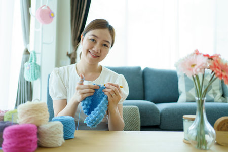Happy cheerful Asian woman doing a crochet in living room in free time. Skillful Asian craftswoman knitting a handcraft crochet sweater and hat in cozy living room.の写真素材