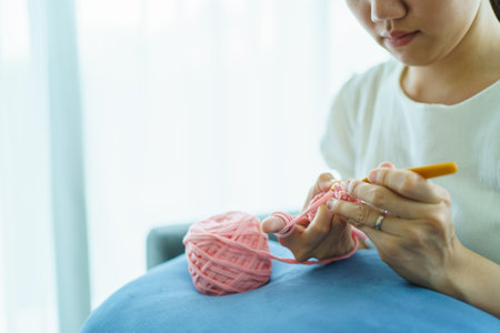 Happy cheerful Asian woman doing a crochet in living room in free time. Skillful Asian craftswoman knitting a handcraft crochet sweater and hat in cozy living room.の写真素材