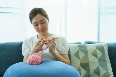 Happy cheerful Asian woman doing a crochet in living room in free time. Skillful Asian craftswoman knitting a handcraft crochet sweater and hat in cozy living room.の写真素材