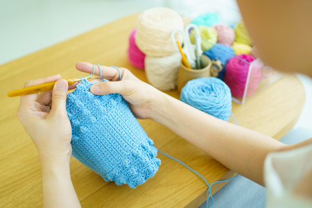 Happy cheerful Asian woman doing a crochet in living room in free time. Skillful Asian craftswoman knitting a handcraft crochet sweater and hat in cozy living room.の写真素材