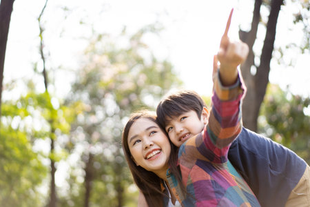 Asian woman and boy playing and walking around the park together with beautiful bokeh and lighting background copyspace. Little cute boy hogging his mother from back and kissing.の写真素材
