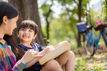 Happy cheerful Asian lovely little boy and mother sitting under the tree at park in the morning and drawing a picture. Asian woman spending time in weekend with her lovely little son.の写真素材