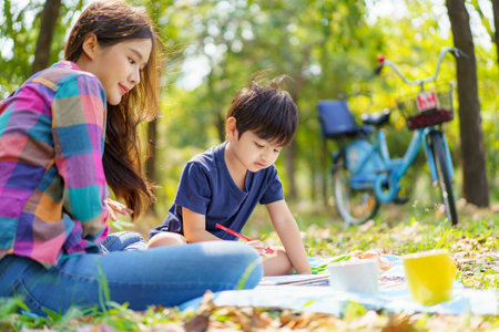 Happy cheerful Asian lovely little boy and mother sitting under the tree at park in the morning and drawing a picture. Asian woman spending time in weekend with her lovely little son.の写真素材