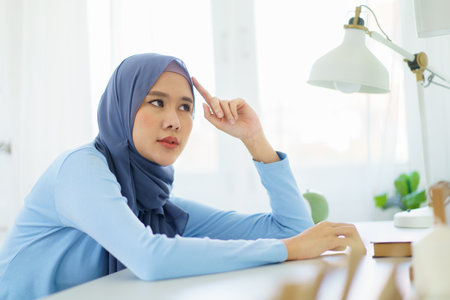 Asian young female muslim student feels sleepy and yawn while doing her homework at home. Lazy - bored muslim woman sitting in the home office and yawning.の写真素材