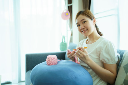 Happy cheerful Asian woman doing a crochet in living room in free time. Skillful Asian craftswoman knitting a handcraft crochet sweater and hat in cozy living room.の写真素材
