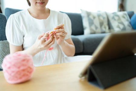 Happy cheerful Asian woman doing a crochet in living room in free time. Skillful Asian craftswoman knitting a handcraft crochet sweater and hat in cozy living room.の写真素材