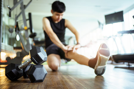 Happy cheerful Asian sportsman relaxing - sitting on the floor after done a weight training exercise in an indoor fitness or gym. Man resting after did a wight training workout.の写真素材