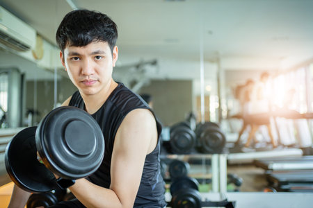 Happy cheerful Asian sportsman doing a weight training exercise at bicep muscular by weight lifting. Asian sporty man doing a dumbbell weight lifting workout in an indoor fitness gym.の写真素材