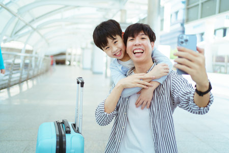Happy cheerful Asian man and little boy traveling on the city railway station and using smartphone to take a selfie photography. Father and son using smartphone to take a photo or video call.の写真素材