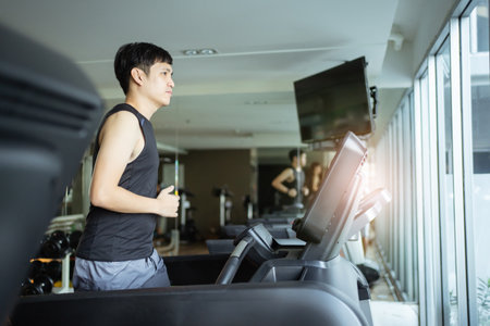 Active Asian sportsman making a cardio exercise by running on the treadmill machine in a fitness or...