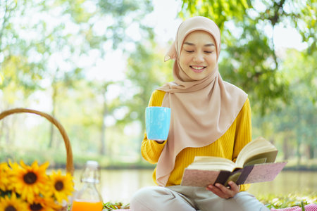 Happy cheerful Asian beautiful muslim woman sitting on the ground at the park and drinking a cup of tea in afternoon. Beautiful muslim woman relaxing and picnic in weekend.の写真素材