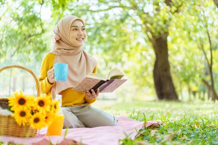 Happy cheerful Asian beautiful muslim woman sitting on the ground at the park and drinking a cup of tea in afternoon. Beautiful muslim woman relaxing and picnic in weekend.の写真素材