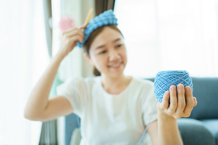 Happy cheerful Asian woman doing a crochet in living room in free time. Skillful Asian craftswoman knitting a handcraft crochet sweater and hat in cozy living room.の写真素材