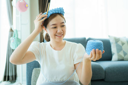 Happy cheerful Asian woman doing a crochet in living room in free time. Skillful Asian craftswoman knitting a handcraft crochet sweater and hat in cozy living room.の写真素材