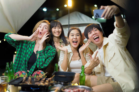 Group of diverse ethnicity young people enjoy eating and grilling bar-b-q together while camping in forest at night. Happy friends enjoy drinking a beer and playing a guitar between camping.の写真素材