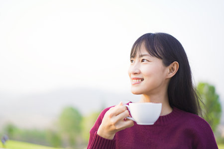 Happy beautiful Asian young woman sitting on the grass field and enjoy relaxing with a stunning view of mountain and forest landscape. Asian young woman drinking a cup of coffee in morning.の写真素材