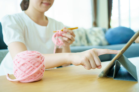 Happy cheerful Asian woman doing a crochet in living room in free time. Skillful Asian craftswoman knitting a handcraft crochet sweater and hat in cozy living room.の写真素材