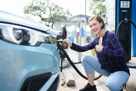 Happy beautiful caucasian white woman driver preparing to recharge her electric vehicle or EV car at EV fast charge station during the trip. Future and sustainable transportation concept.の写真素材