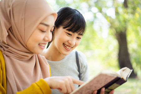Happy beautiful Asian females Muslim student and friends reading a textbook together in a park. Diversity in ethnic and religious concept.の写真素材
