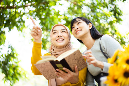Happy beautiful Asian females Muslim student and friends reading a textbook together in a park. Diversity in ethnic and religious concept.の写真素材