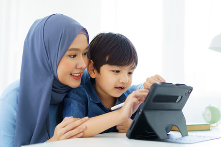 Happy cheerful Asian muslim woman woman teaching her little son a drawing on digital tablet, mother let her little young boy doing a school homework or an online learning on tablet.の写真素材