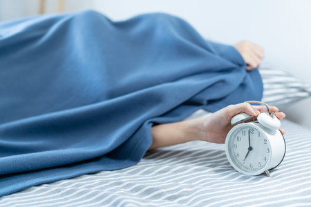 Asian young woman sleeping on the cozy bed in the early morning and trying to turn off or snooze an alarm clock. Lazy sleepy woman wake up late in the morning.の写真素材