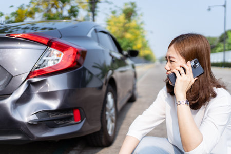 Upset unhappy Asian woman have a car accident or crashed during her road trip, woman driver sitting beside the crashed or broken car and using cell phone calling to road side assistant service.の写真素材