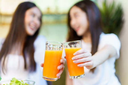 Asian women enjoying eating a breakfast and drinking organic orange juice together in the dining room. Cute Asian women cheering an orange juice in a glass together.の写真素材