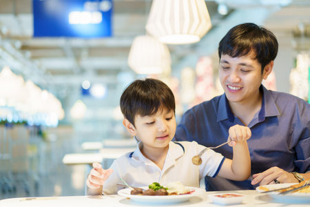 Happy cheerful Asian man and lovely little son enjoy eating a food in the fast food restaurant together. Man looking his son eating a meat ball.の写真素材