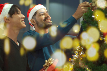 Lovely gay couple enjoy decorating a Christmas tree together in a living room with beautiful festive light bulbs and presents. LGBTQ gay couple celebrating a Christmas Eve and New Year festival.の写真素材