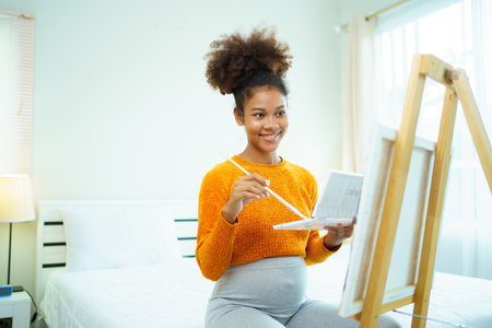 Happy cheerful American - African black ethnicity pregnant woman enjoy drawing and painting a water color on canvas in bedroom, hobby and activity for pregnant woman.の写真素材