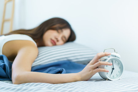 Asian young woman sleeping on the cozy bed in the early morning and trying to turn off or snooze an alarm clock. Lazy sleepy woman wake up late in the morning.の写真素材
