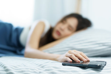 Asian young woman got a phone call on smartphone while sleeping on the bed in morning and trying to ignore incoming phone call. Woman turn off or snooze his alarm clock on smartphone.の写真素材