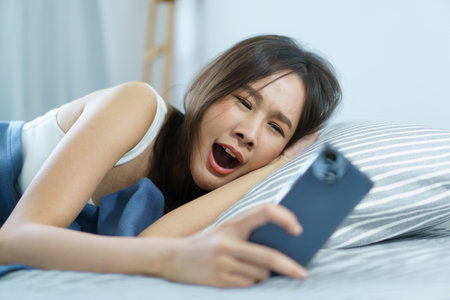 Asian young woman got a phone call on smartphone while sleeping on the bed in morning and trying to ignore incoming phone call. Woman turn off or snooze his alarm clock on smartphone.の写真素材