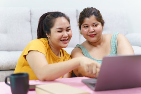 Happy Asian women friend relaxing in living room and drink a cup of tea together. Happy - delighted two Asian oversized women talking and watching a movie on laptop or notebook computer.の写真素材