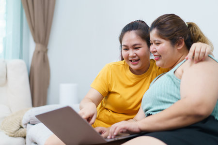 Happy Asian women friend relaxing in living room and drink a cup of tea together. Happy - delighted two Asian oversized women talking and watching a movie on laptop or notebook computer.の写真素材