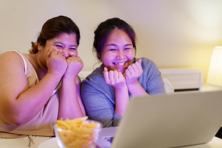Asian oversize fat women sitting on the bed and enjoy eating French Fry and watching movie on laptop - notebook computer at night. Oversize fat woman watching a movie or TV show steaming on laptop.の写真素材