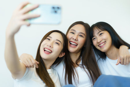Happy beautiful Asian young women staying together in bedroom and taking a selfie photography on smart phone, woman friends waving hands while taking a selfie photo on mobile phone.の写真素材
