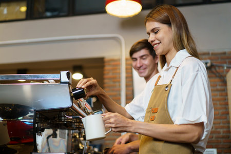 Professional Asian female barista pouring a hot milk over the espresso in coffee cup. Happy female barista making a cup of latte coffee in a small coffee shop. Small business concept.の写真素材
