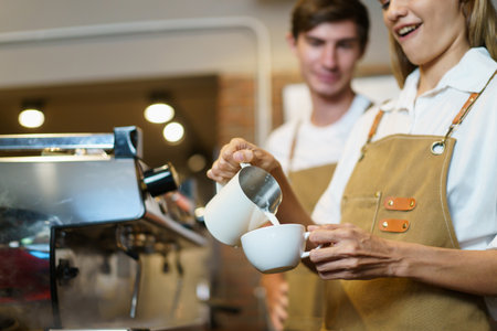 Professional Asian female barista pouring a hot milk over the espresso in coffee cup. Happy female barista making a cup of latte coffee in a small coffee shop. Small business concept.の写真素材
