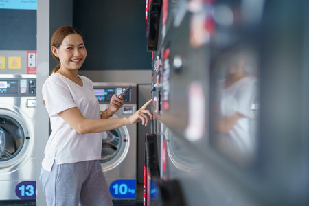 Happy Asian beautiful woman using a self service washing machine KIOSK or automatic washing machine vendor. Woman using smartphone control the washing machine.の写真素材