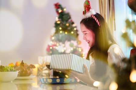 A beautiful Asian woman is excited to open Christmas presents during her Christmas dinner in a cozy and warm living room at home. She is enjoying celebrating Christmas night alone.の写真素材